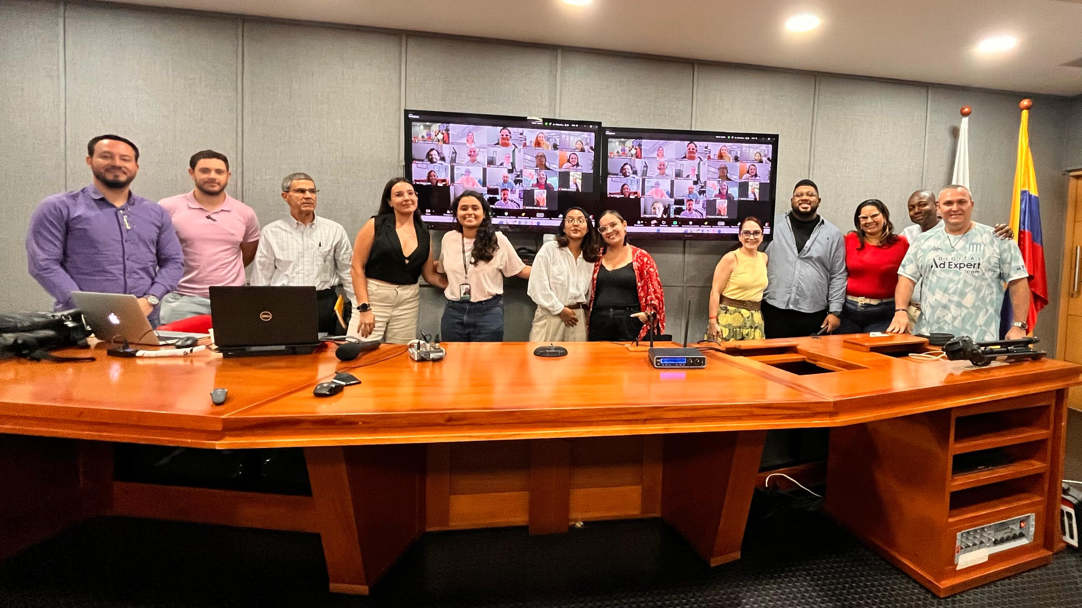 Descripción de imagen: se aprecia a un grupo de personas, algunas abrazadas y sonriendo, la mayoría de pie y otras en la virtualidad en un auditorio