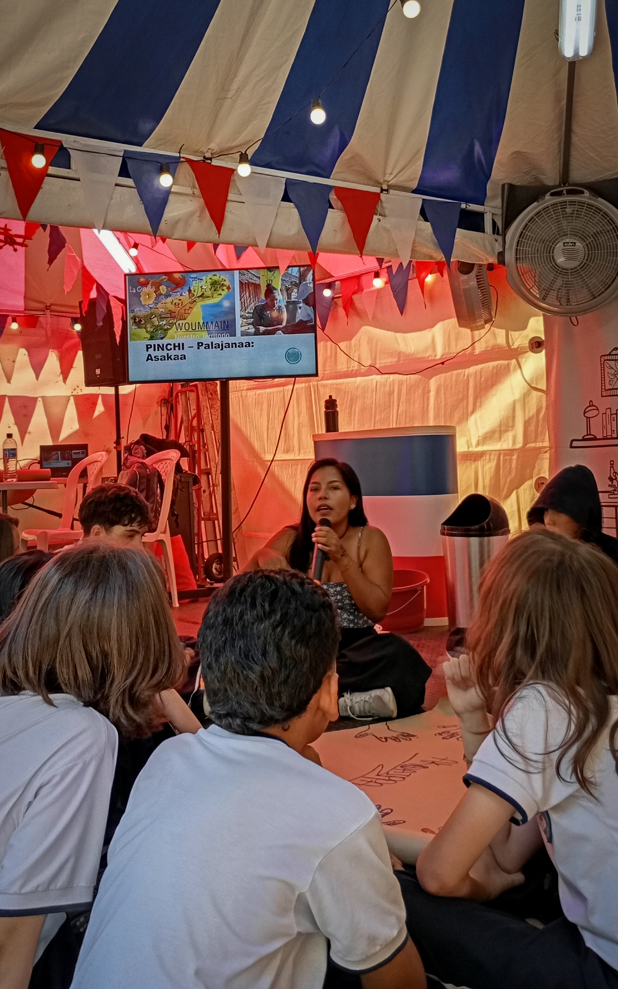 Profesora hablando a estudiantes sentados dentro de una carpa