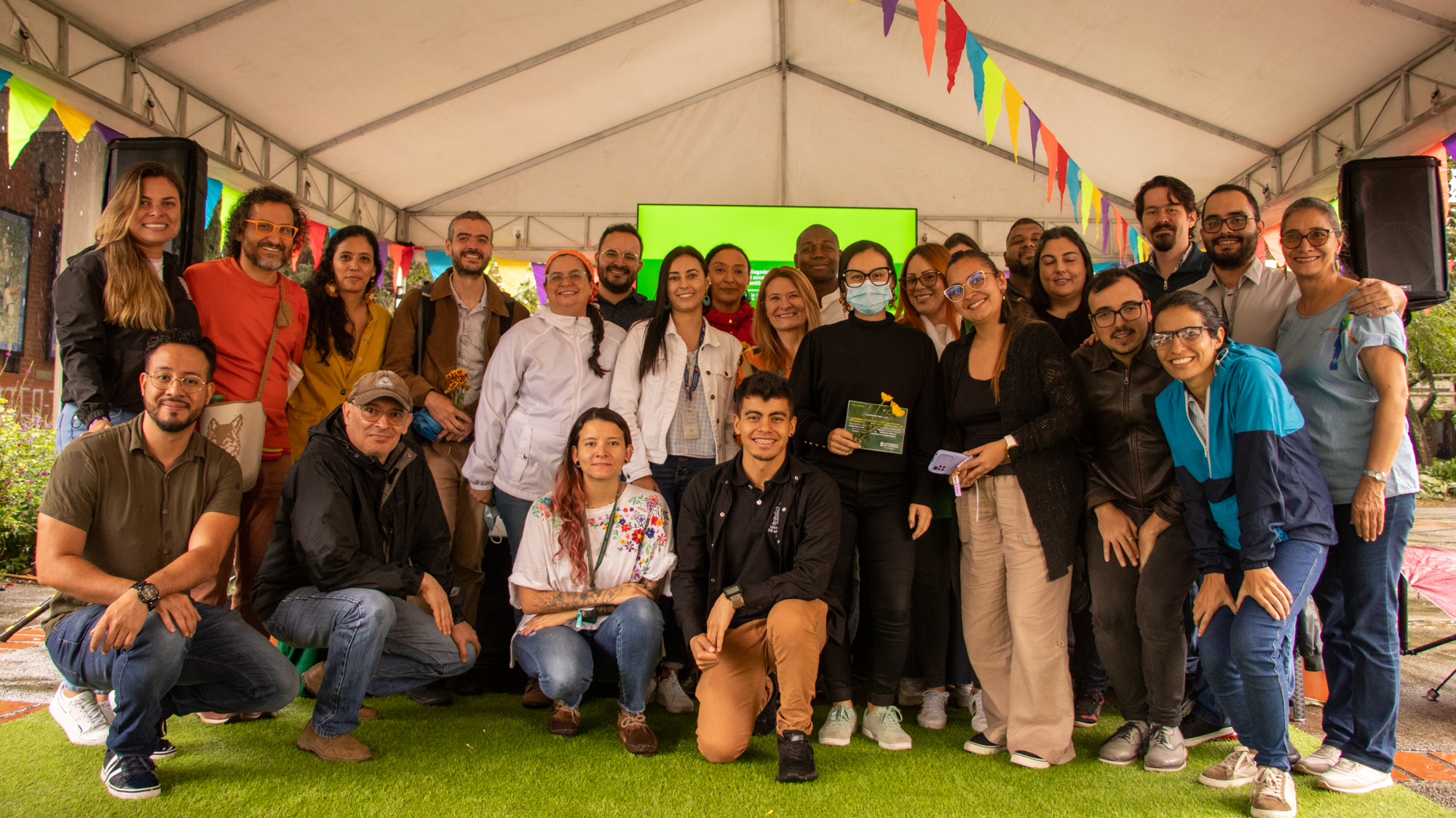 La imagen muestra un grupo diverso de personas mirando sonrientes a la cámara reunidas bajo una carpa decorada con banderines de colores en Ciudadela Universitaria.