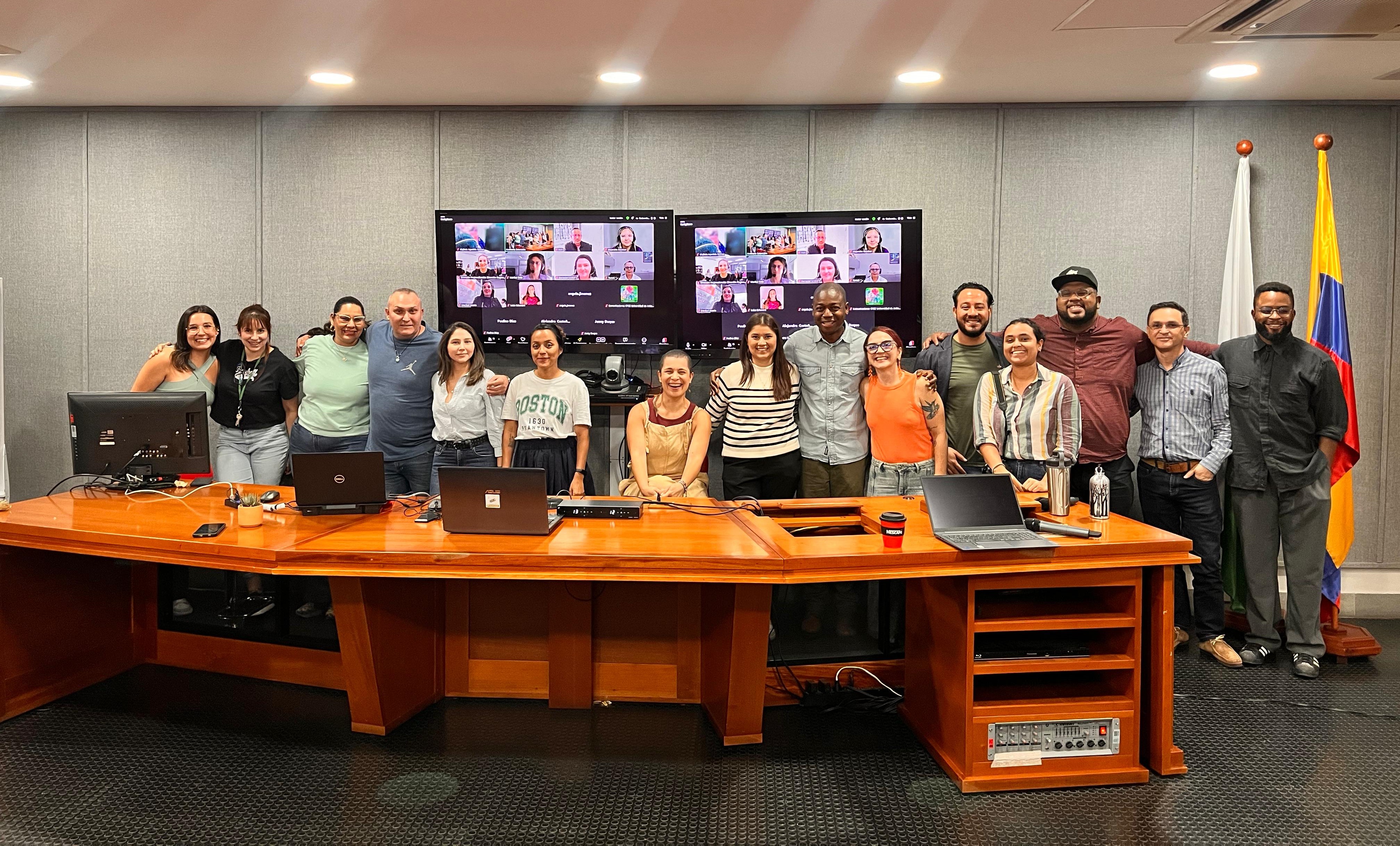 Descripción de imagen: se aprecia a un grupo de personas, algunas abrazadas y sonriendo, la mayoría de pie y otras en la virtualidad en un auditorio de la reunión del 5 de marzo.