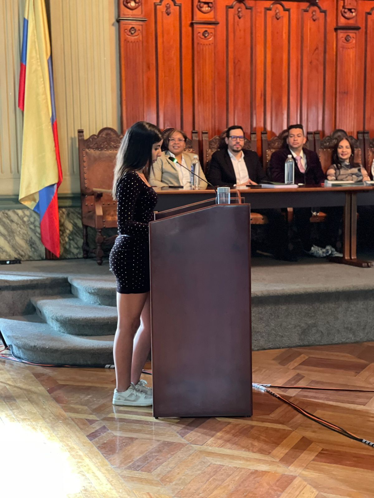 Estudiante de pie en atril. De fondo mesa principal