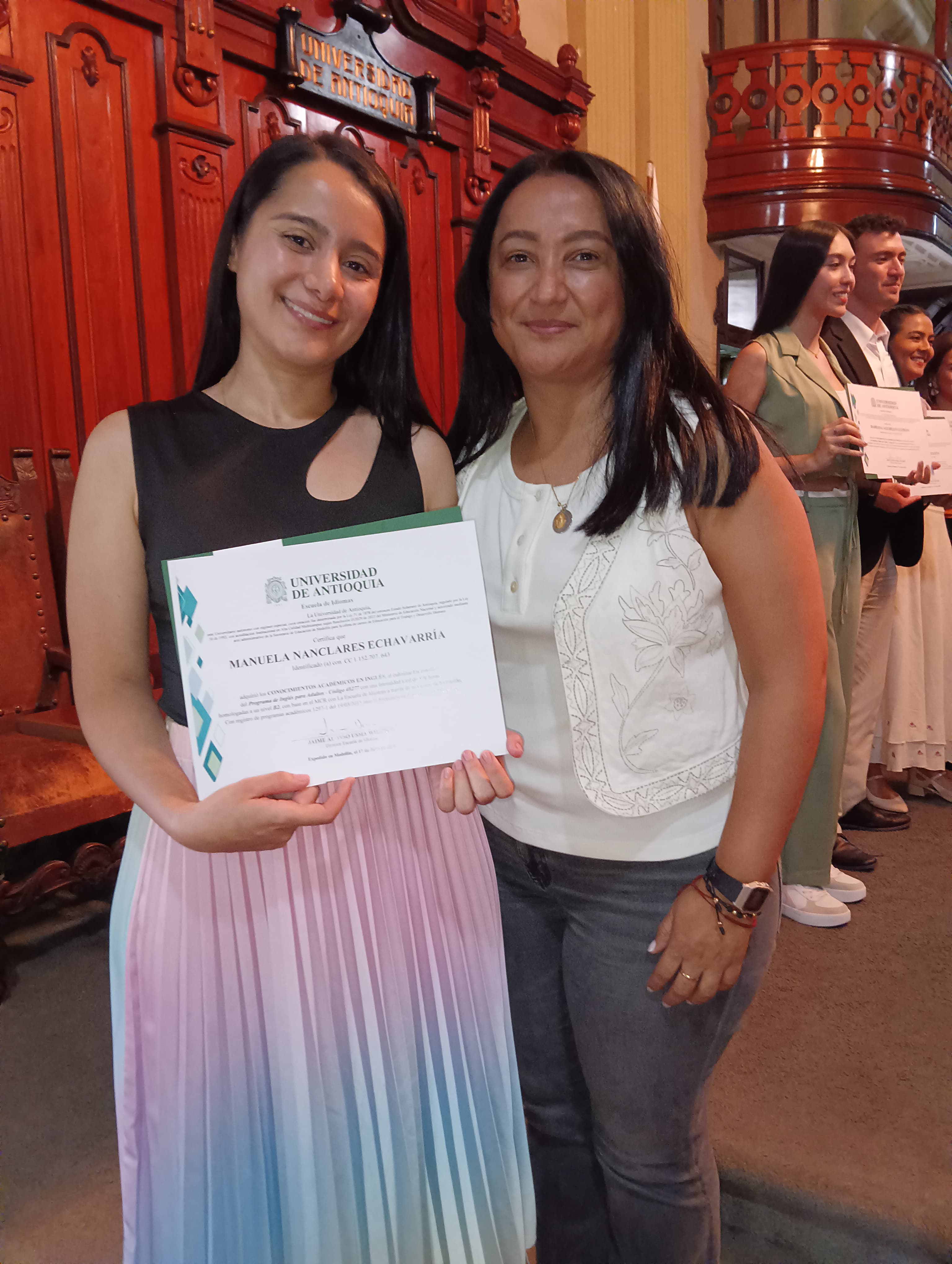 Estudiante de pie con diploma en las manos al lado de su mamá
