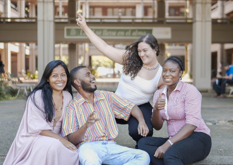 Descripción de imagen: un grupo de mujeres y un hombre de diversas culturas sonríen. Una de las mujeres hace la seña de estar bien con el dedo pulgar hacia arriba. En el fondo aparece el Bloque 16 de la Ciudad Universitaria.