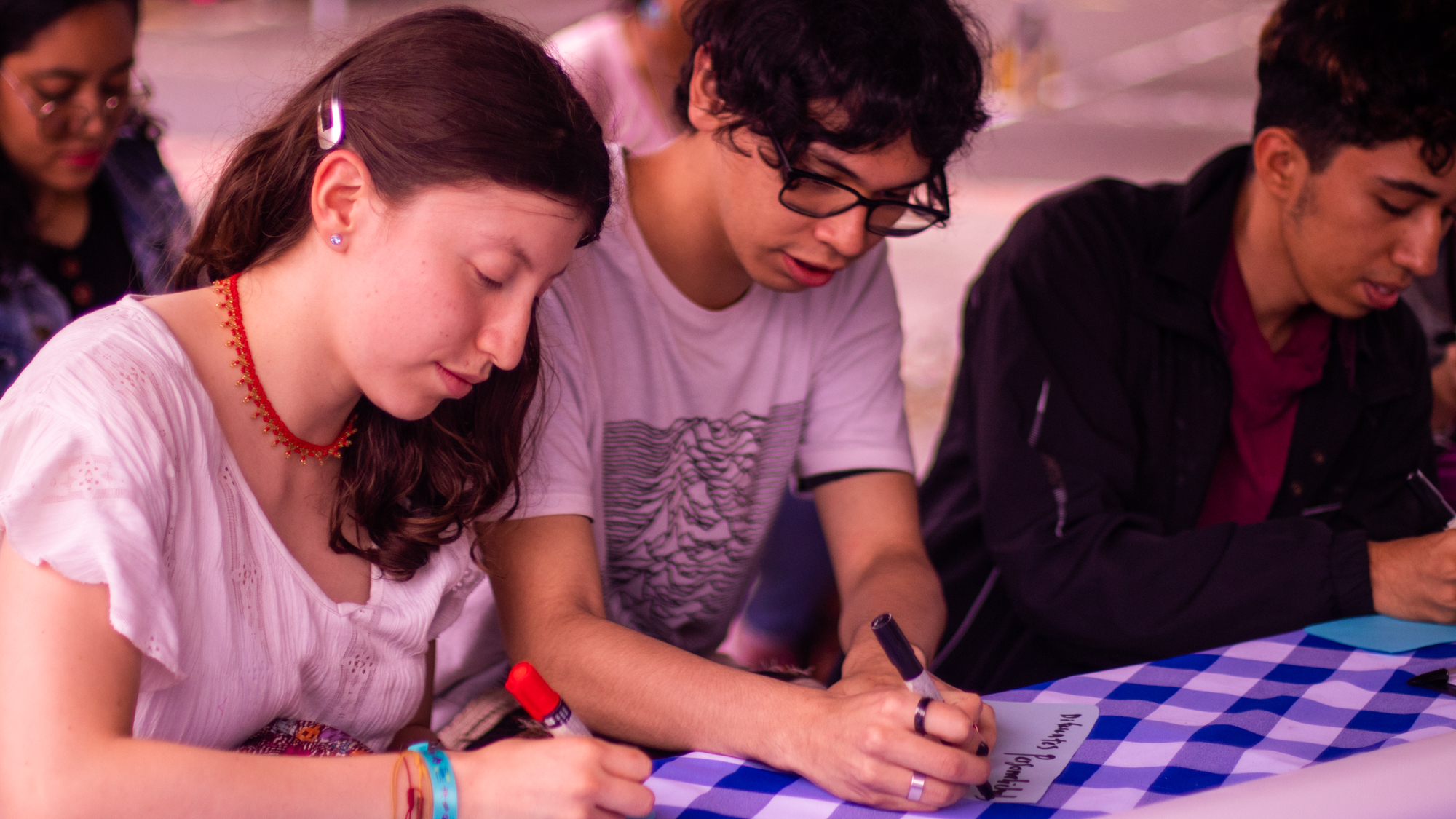 tres jóvenes sentados escribiendo sobre una mesa con mantel de cuadros azules y blancos. Todos parecen concentrados en su tarea.