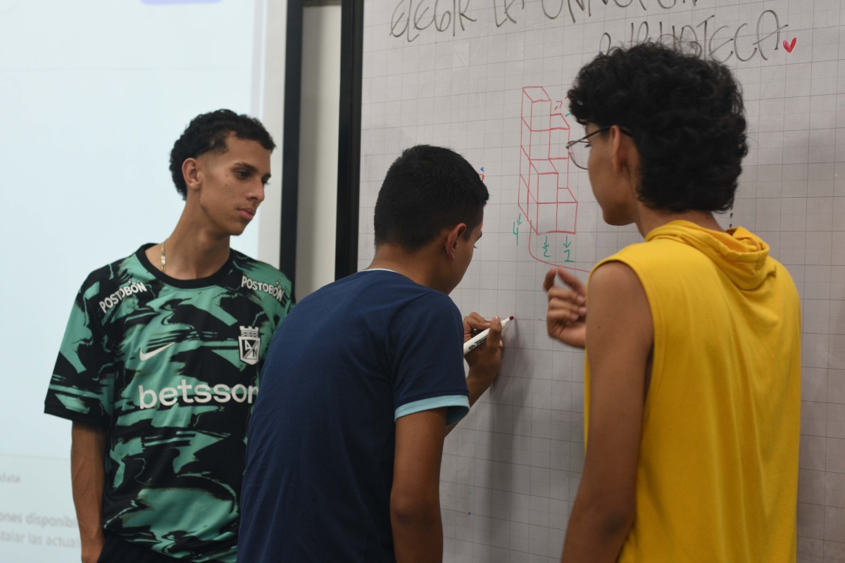 Aparecen tres participantes de «Señando la U» frente a un tablero, trabajando juntos en la resolución de un ejercicio de razonamiento lógico. Uno de ellos escribe con marcador rojo debajo de una figura geométrica.