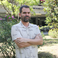 Foto del profesor Héctor Mauricio. El fondo tiene árboles. El tiene una camisa blanca con un patrón en color negro.