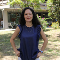 Fotografía de Sara. Ella tiene una camisa azul y un pantalón blanco. Al fondo se ve la naturaleza y la parte de atrás del Bloque 13 Fotografía de Sara. Ella tiene una camisa azul y un pantalón blanco. Al fondo se ve la naturaleza y la parte de atrás del Bloque 13