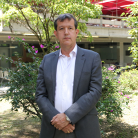 Foto del Profesor Jairo Humberto. El fondo tiene árboles. El tiene un traje gris y una camisa rosa Foto del Profesor Jairo Humberto. El fondo tiene árboles. El tiene un traje gris y una camisa rosa